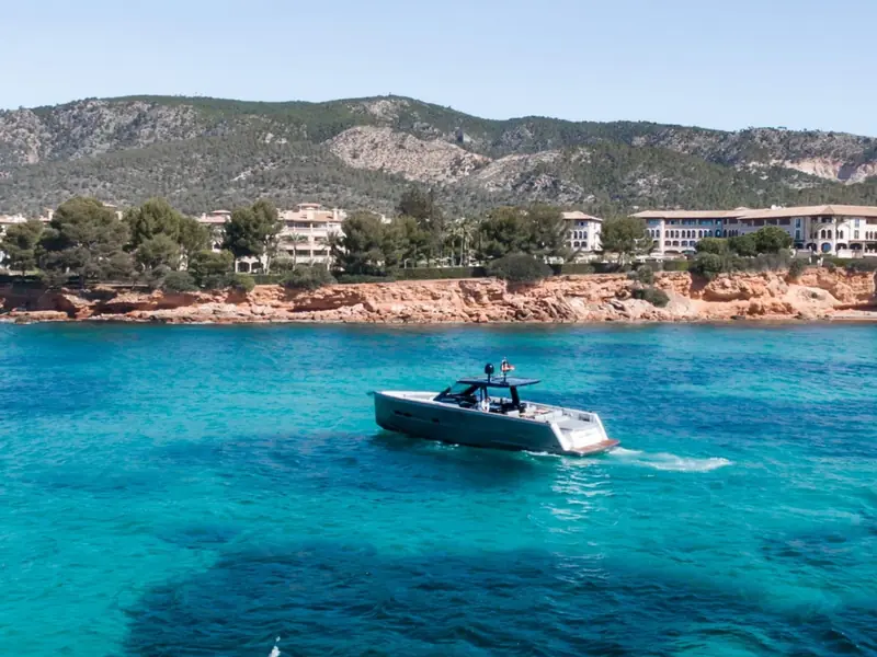 BOMBON, a Fjord 48 Open motor yacht, cruising in clear turquoise water near a rocky coast with buildings.