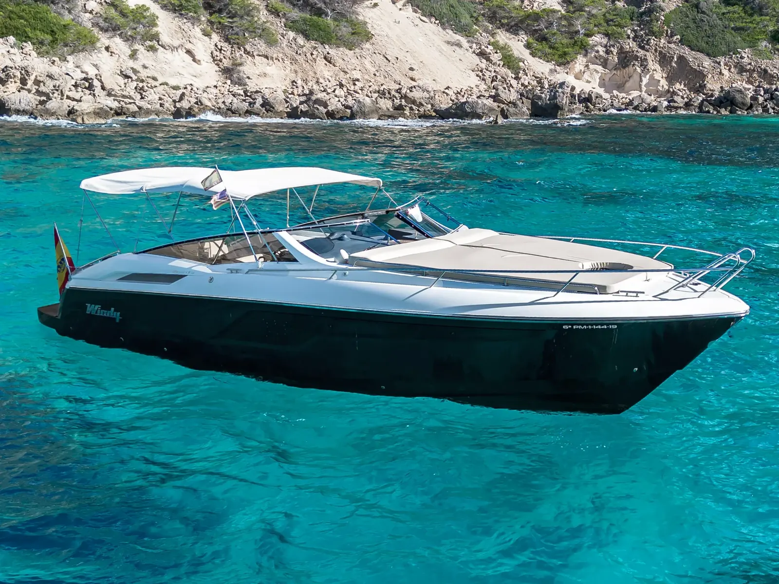 CUCA, a Windy 39 Camira with a dark hull and white bimini top, anchored in clear turquoise water near rocky cliffs.