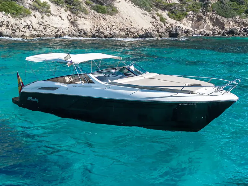 CUCA, a Windy 39 Camira with a dark hull and white bimini top, anchored in clear turquoise water near rocky cliffs.