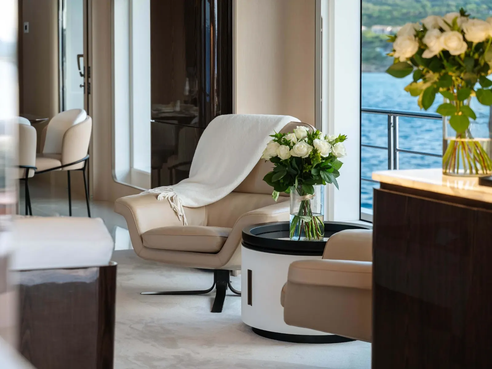 NEMESIS yacht interior showing a beige armchair, white roses, and a view of the water through floor-to-ceiling windows.