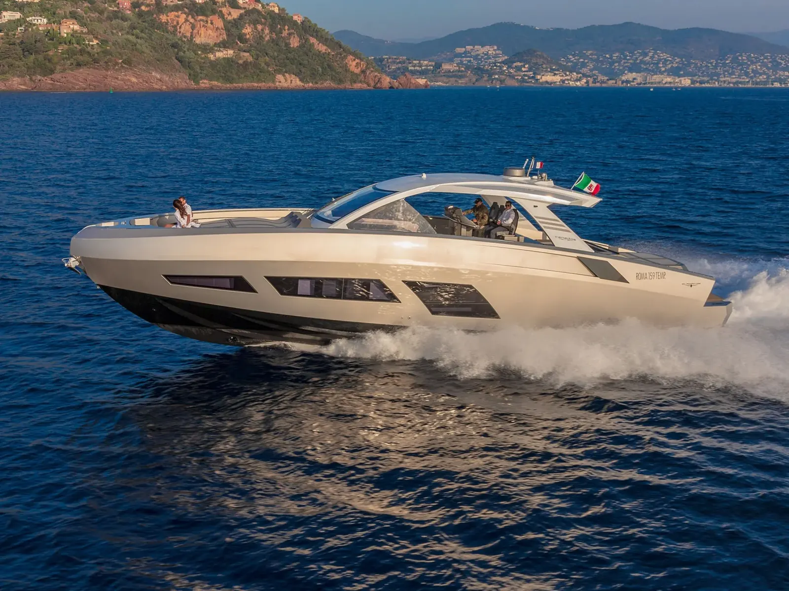 Yacht NEXT, a Canados 631, cruising at speed on blue water near a coastline with hills and buildings.
