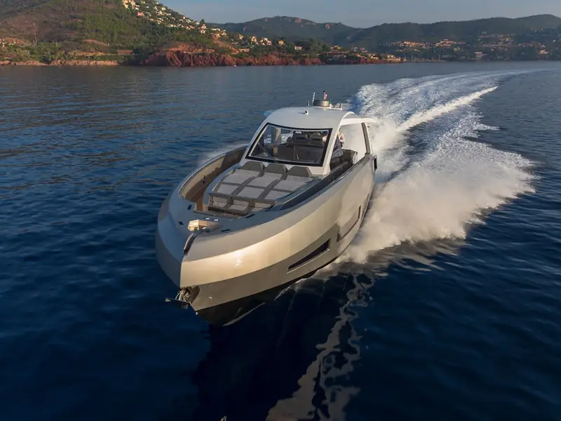 NEXT yacht cruising at speed, showing its grey hull, foredeck sunpads, and white hardtop against a coastal backdrop.