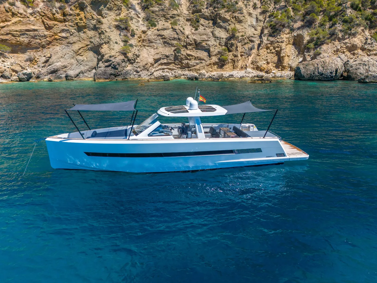 PETITCHET yacht anchored near rocky cliffs with sunshades deployed over the foredeck and aft deck.