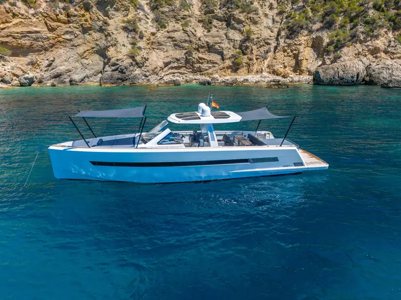 PETITCHET yacht anchored near rocky cliffs with sunshades deployed over the foredeck and aft deck.