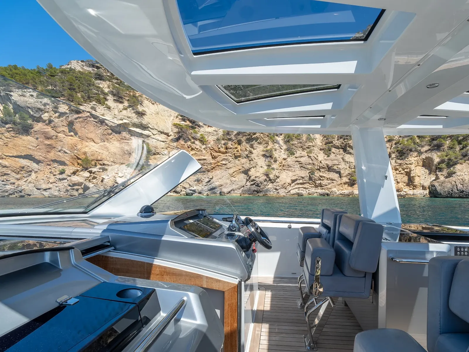 PETITCHET helm station with grey seats and glass roof panels, anchored near a rocky cliff.