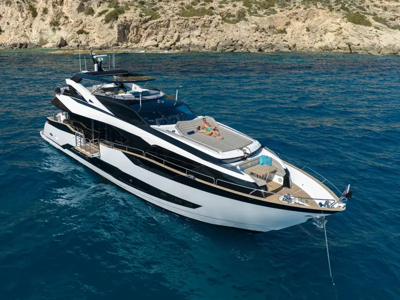 BLUE INFINITY ONE yacht anchored at sea with people on the foredeck sunpad near a rocky coastline.