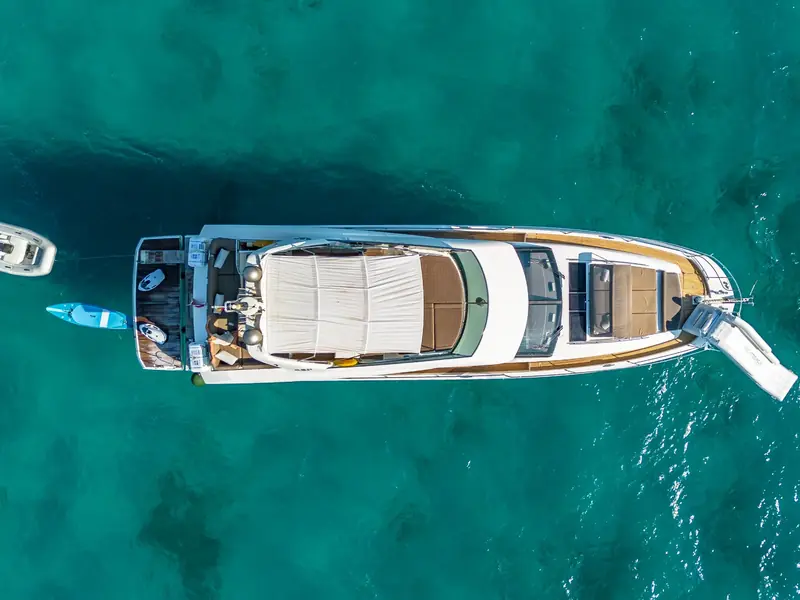 Aerial top-down view of BLUE M in turquoise water with a tender, paddleboard, and inflatable slide deployed.