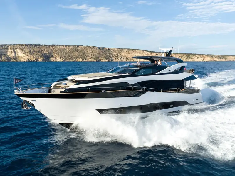 BLUE INFINITY ONE, a Sunseeker 95 yacht, cruising at speed near a rocky coastline.